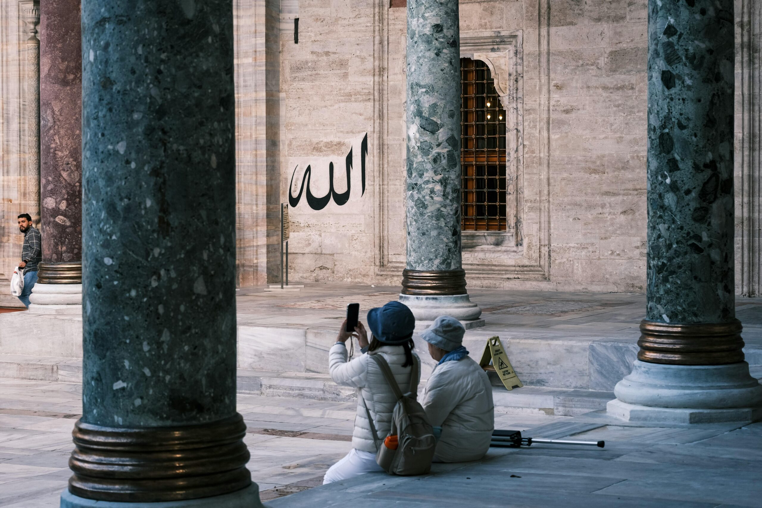 Capture of tourists photographing mosque columns and Arabic calligraphy indoors.